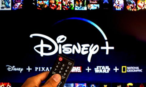 Close-up of a hand holding a remote navigating to Disney Plus on a TV .