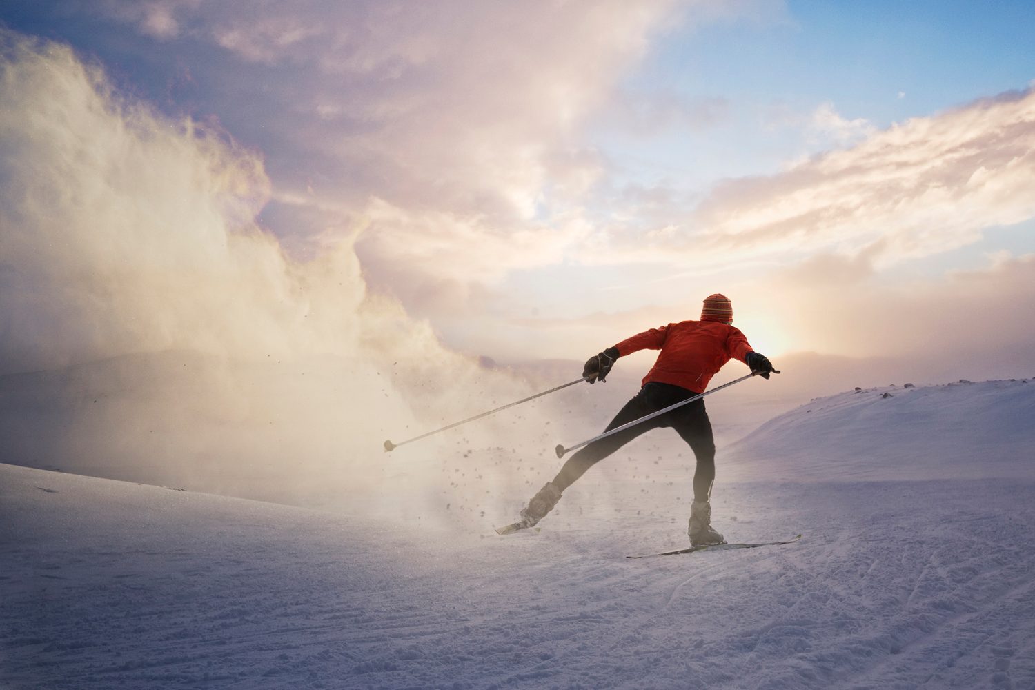 A man cross-country skiing at sunrise, leaving a puff of snow behind his wake.