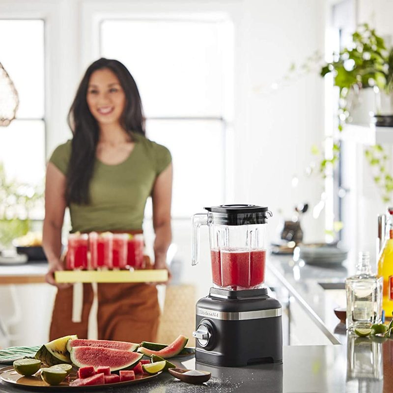 KitchenAid K150 3 Speed Ice Crushing Blender being used by a woman to make smoothies in a home kitchen.