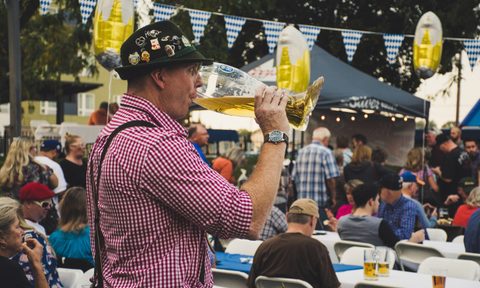 Man dressed in lederhosen drinking beer from a glass boot