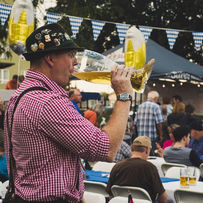man dressed in lederhosen drinking beer from a glass boot.