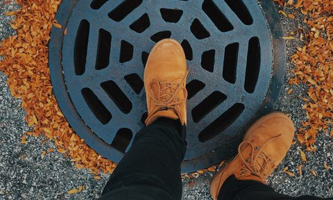 Person wearing Timberland boots while standing on a street drain surrounded by leaves