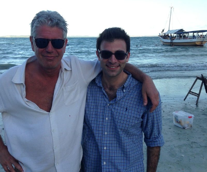 Anthony Bourdain and Tom Vitale standing on a beach together with a boat in the background.