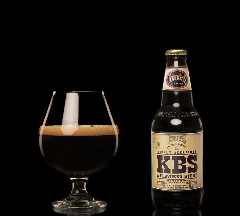 bottle of Founders KBS beer sitting next to a tulip glass of beer against a black background.