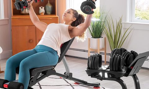 A woman lifting Bowflex SelectTech 552 Adjustable Dumbbells with a Bowflex stand nearby.