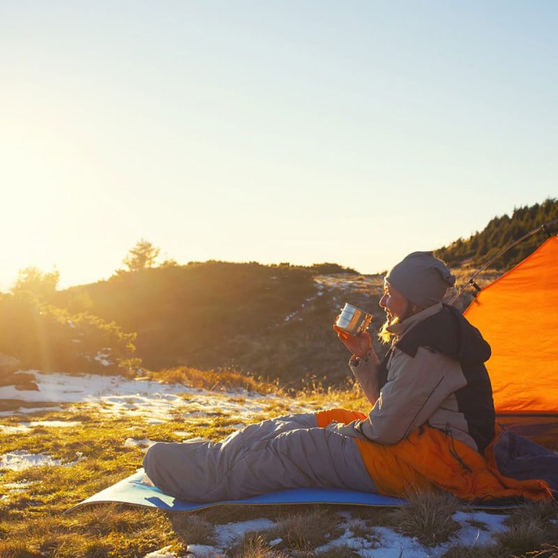 A woman camping outdoors sitting in her sleeping bag and sipping a drink.