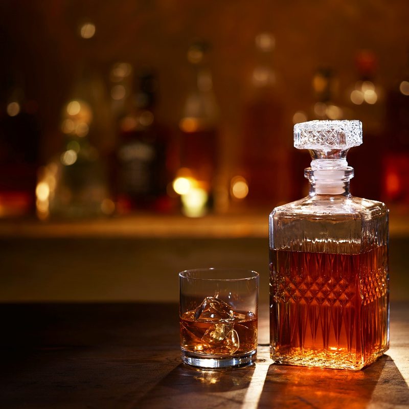 A Bourbon Whiskey Decanter and Bourbon on the Rocks in a Tumbler sitting on a bar with bottles in the background.