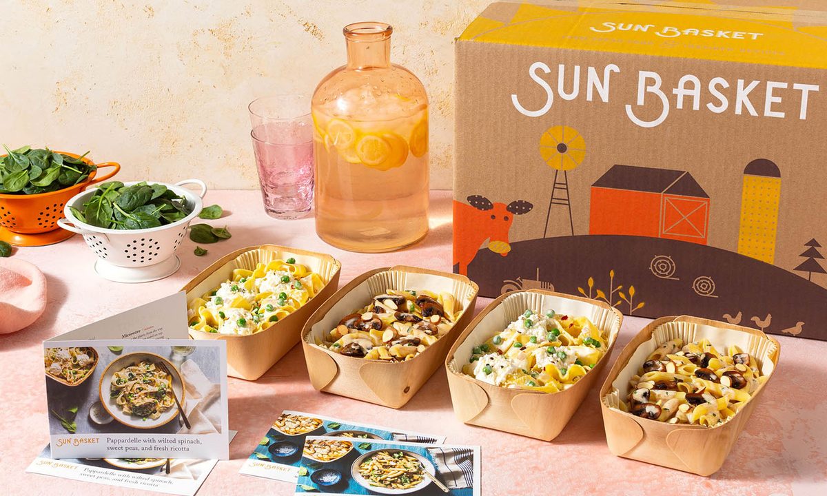 Recipe cards and four sets of healthy meals in front of a Sun Basket box with flavored water and veggie leaves beside them