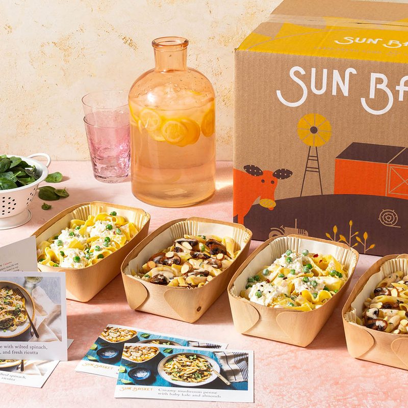 Recipe cards and four sets of healthy meals in front of a Sun Basket box with flavored water and veggie leaves beside them