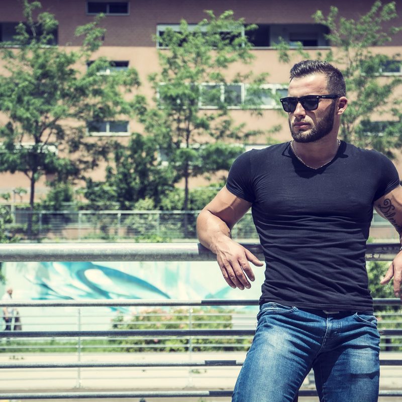 A muscular man in a black T-shirt and blue jeans leaning against a fence.