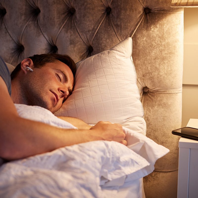 Man Sleeping In Bed Wearing Wireless Earphones Connected To Mobile Phone On Bedside Table.