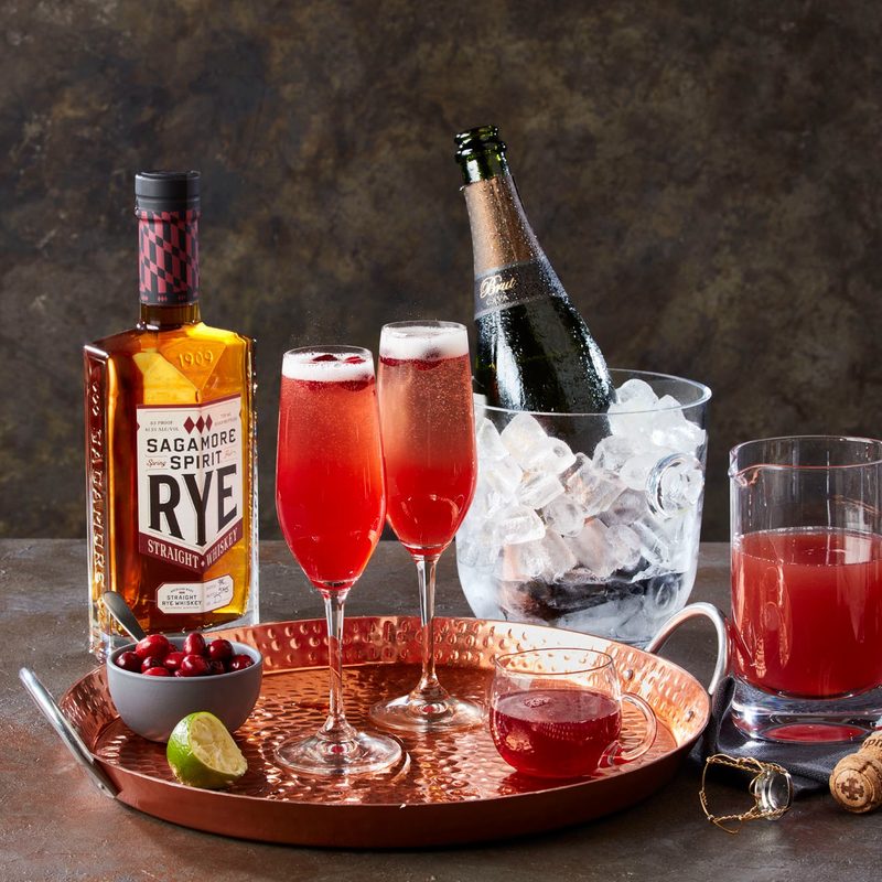 bar setting of two Cava and Cranberry cocktails in champagne flutes next to a bottle of rye and cava on ice.