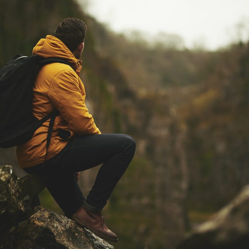 A hiker in a yellow waterproof sits on a rock and looks uphill on an overcast day