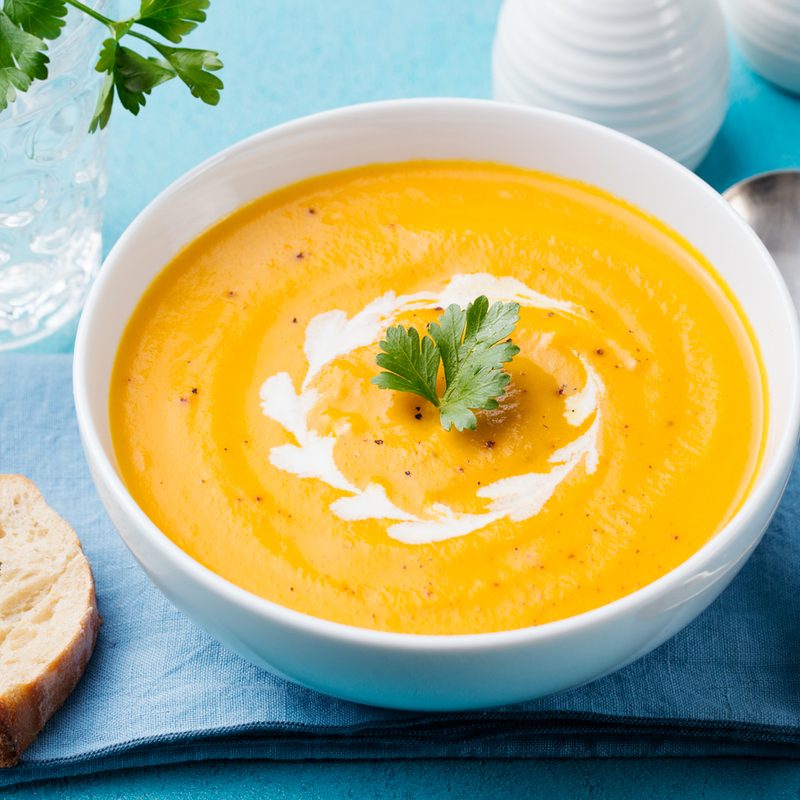 A bowl of carrot purée with parsley beside a slice of bread and a metal spoon.