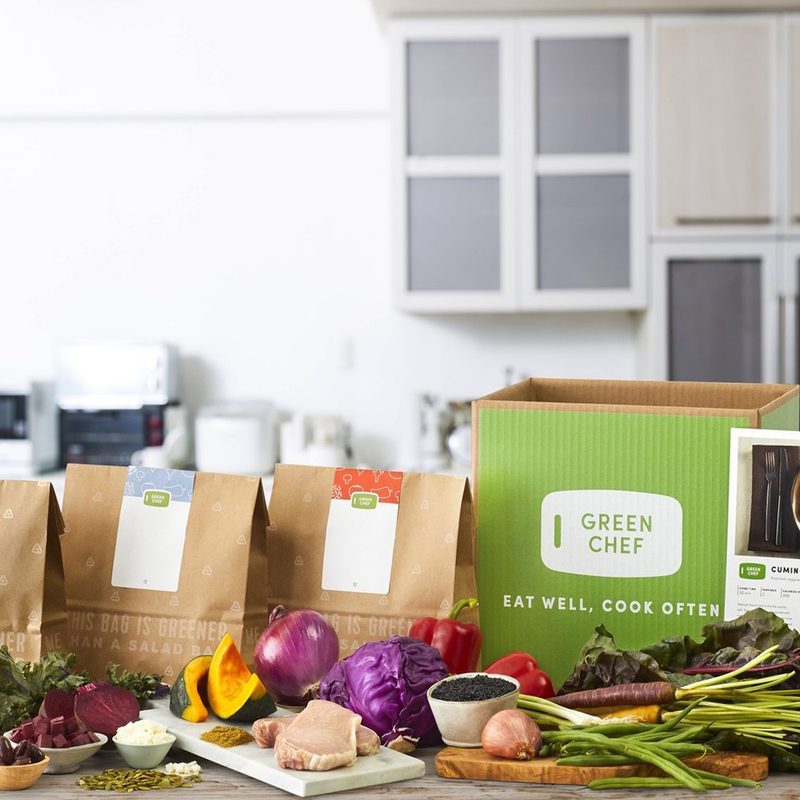 Three paper bags and an empty Green Chef box placed behind a variety of healthy foods on a kitchen counter.