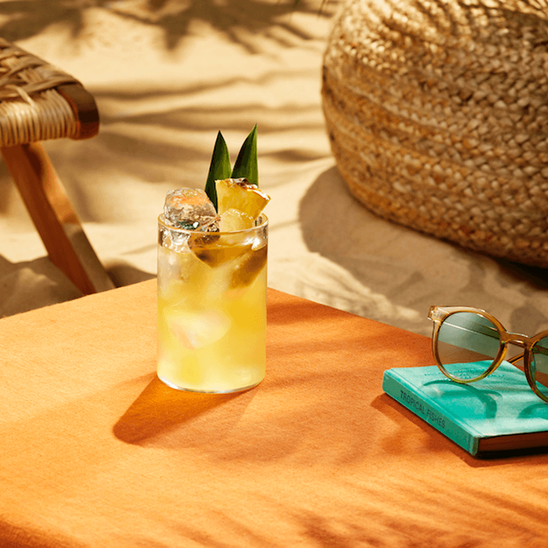 A glass of Life's a Beach cocktail beside a book and sunglasses on a beach table.