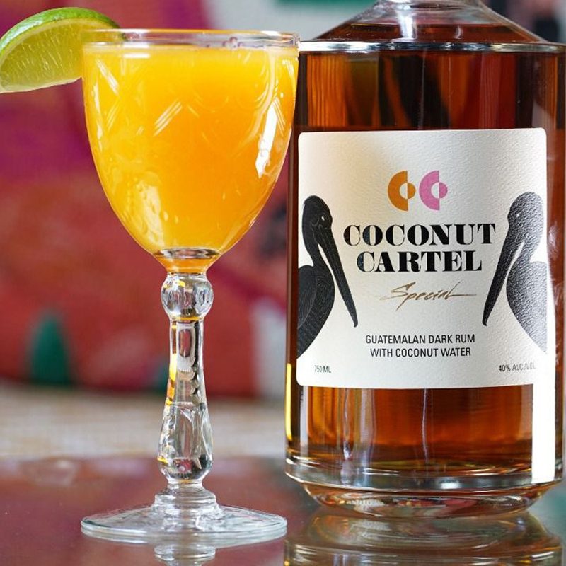 A glass of Little Island Daiquiri beside a bottle of Coconut Cartel.
