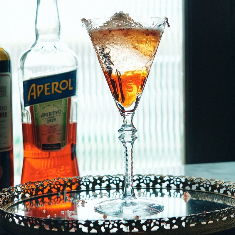 bottle of liquor and aperol sitting next to a martini glass on a mirrored serving tray.