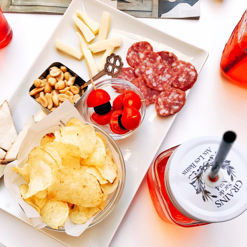 Small plate of cheese, eat, pickled vegetables, and potato chips sitting next to red drinks.