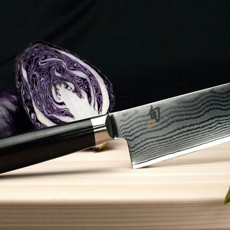 A Shun knife with a sliced red cabbage behind it on a wooden cutting board.