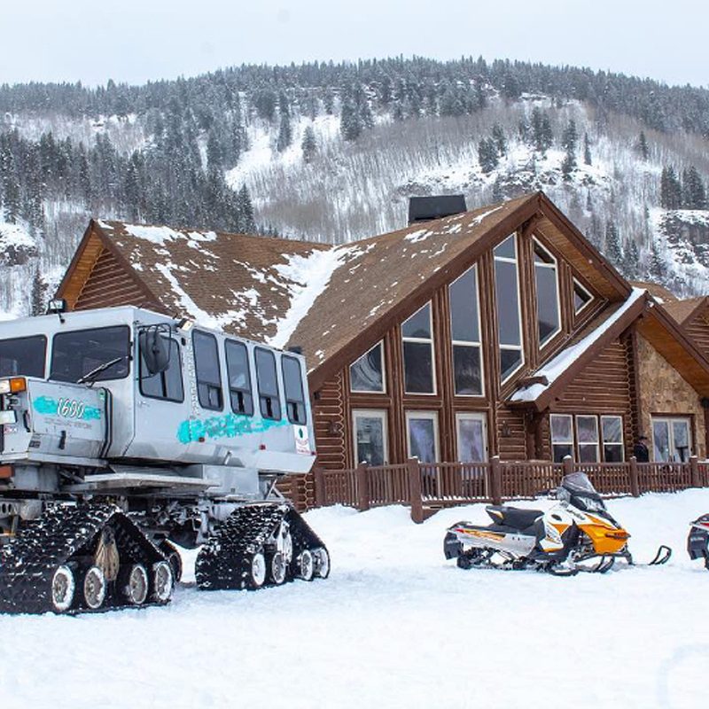 A snowcat and two snowmobiles in front of a cabin.