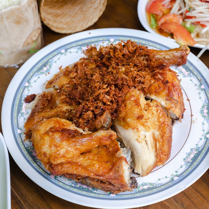 Thai Gai Yang grilled chicken on a plate with som tum salad in the background.
