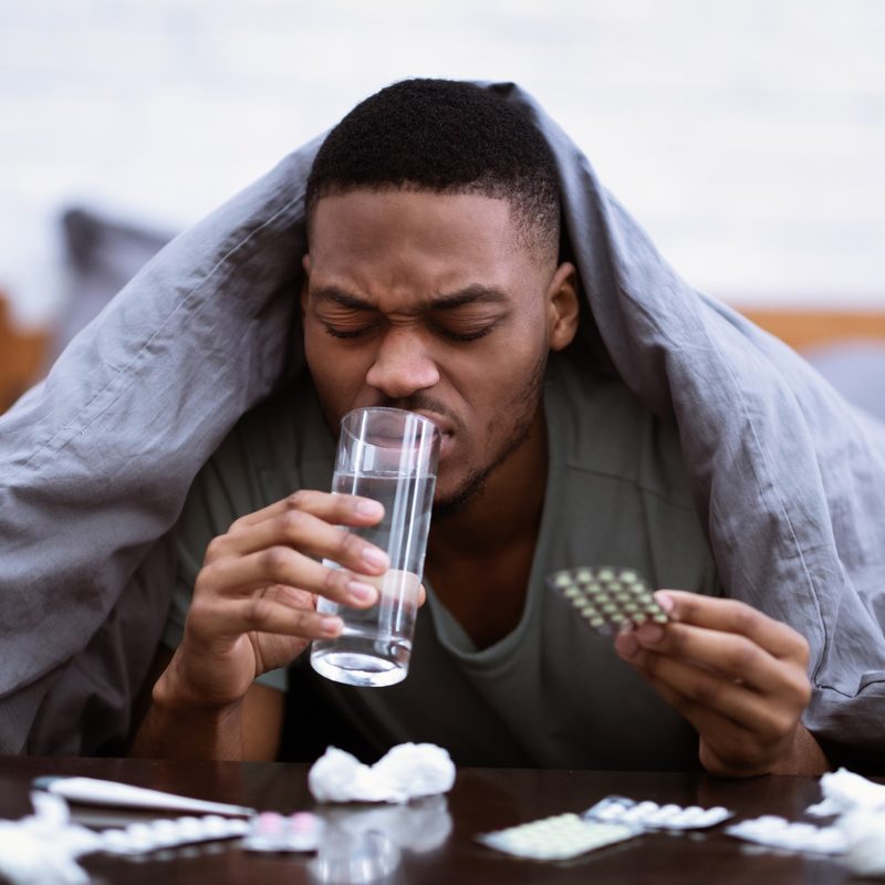Sick man covered in a blanket on a bet, drinking water and taking cold medicine