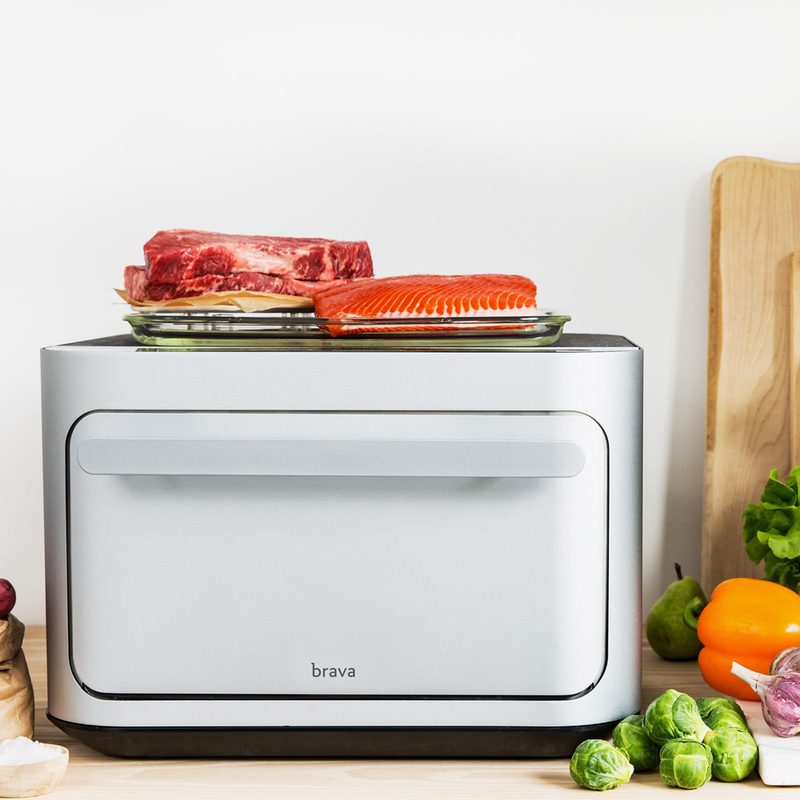 A Brava oven with a tray of fresh salmon slice and meat slabs on top of it, surrounded by healthy foods.