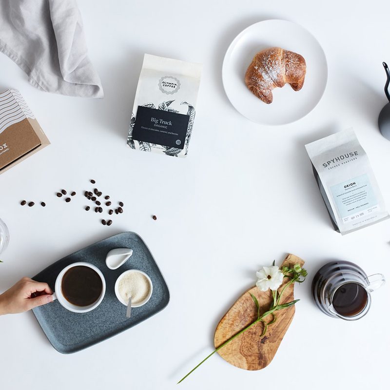 An arm holding a cup of coffee on a tray, surrounded by a box from Mistobox and other items on a table.