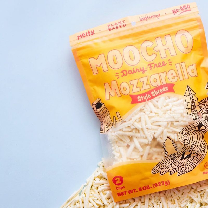 A bag of Moocho Mozarella Style shreds on a blue surface and shredded cheese.