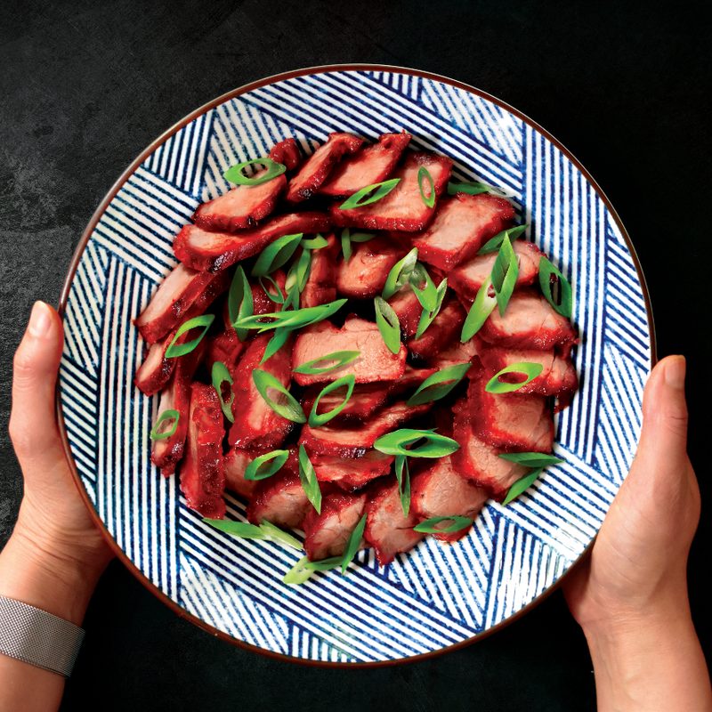 Hands holding a plate of Nom Nom Char Siu, black table in the back.