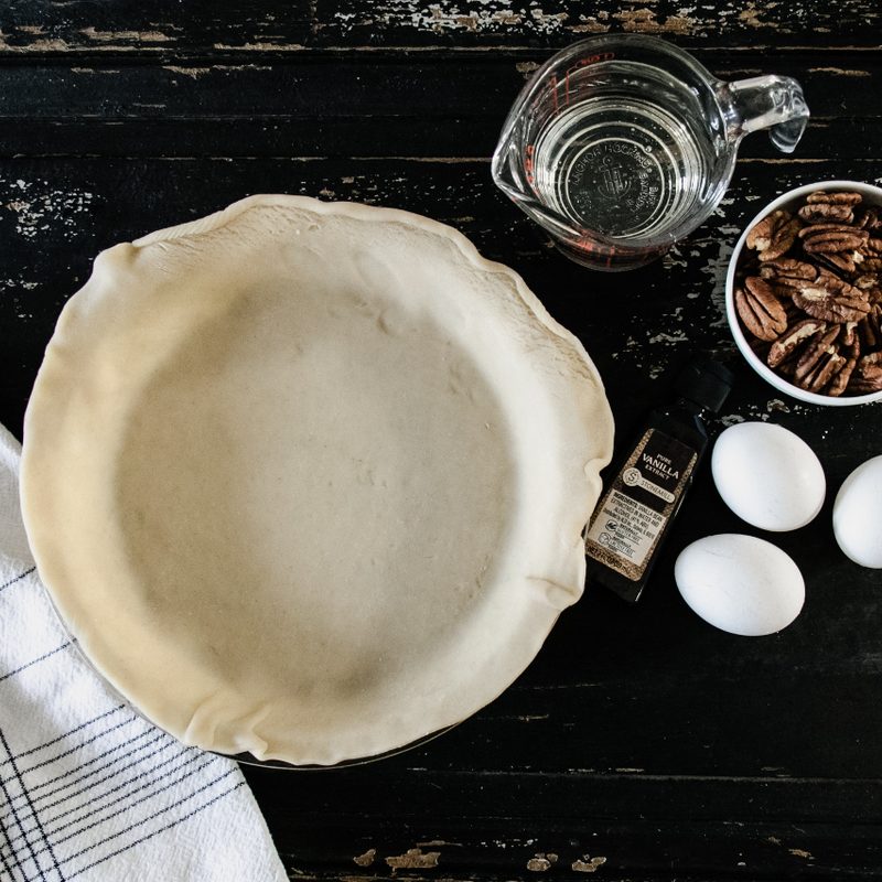 Unfilled pie crusts, 3 eggs, pecans and cup of water on a table.