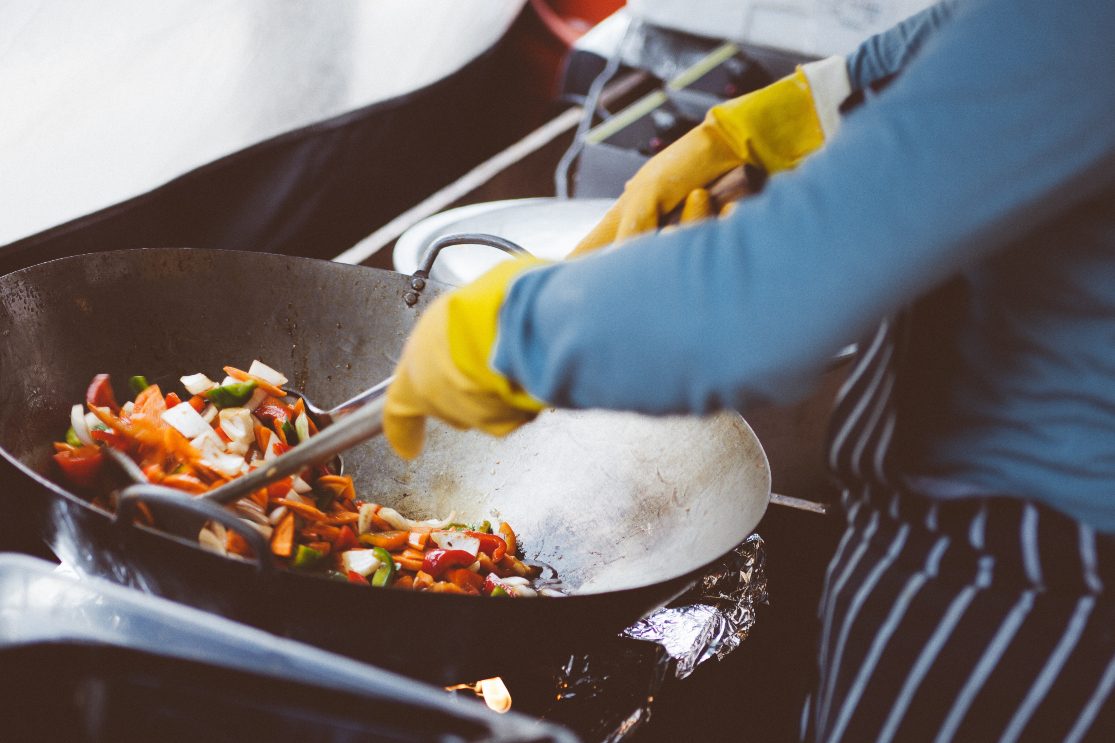 Cook stir frying veggie in a wok wearing yellow gloves.