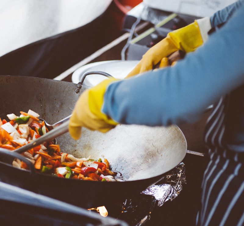 Cook stir frying veggie in a wok wearing yellow gloves.