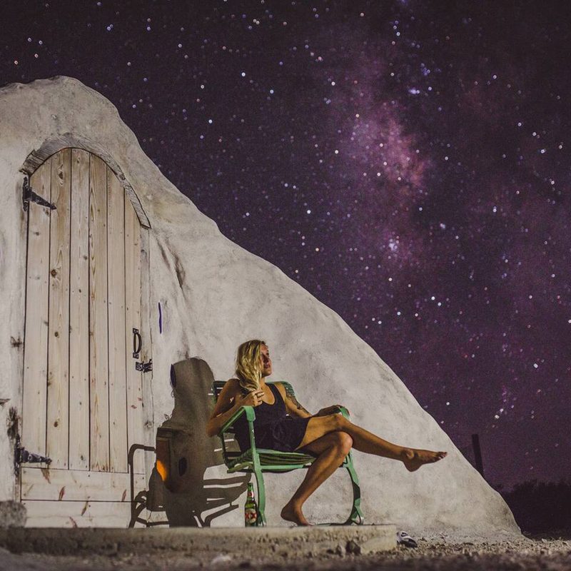 A woman sitting on a bench outside the Terluna Off-Grid Adobe Dome at night.