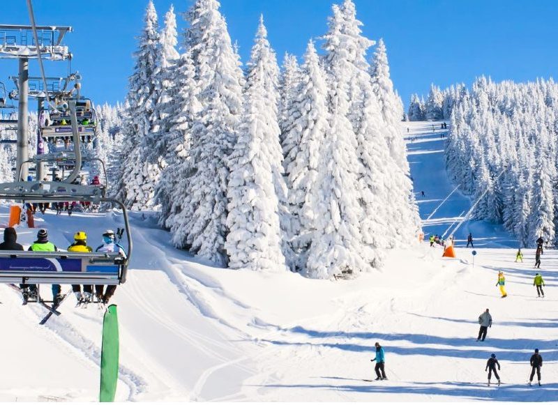 Panorama of ski resort, slope, people on the ski lift, skiers on the piste among white snow pine trees