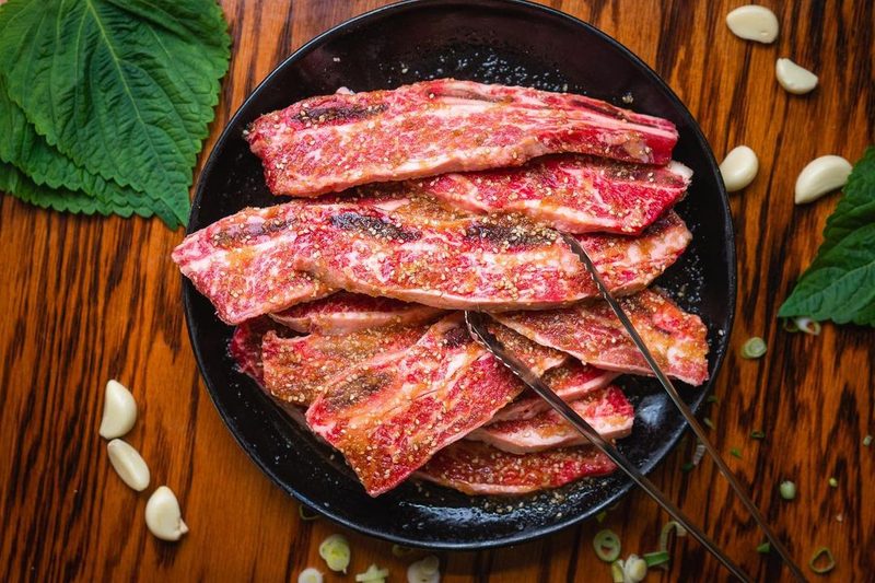 Marinated Korean beef short ribs on a black plate with raw garlic and perilla leaves scattered around.