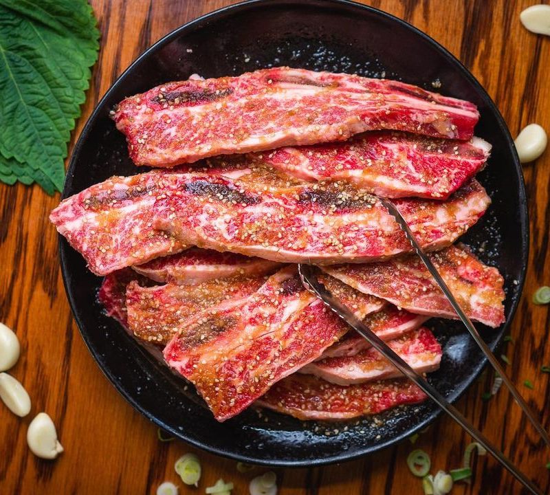 Marinated Korean beef short ribs on a black plate with raw garlic and perilla leaves scattered around.