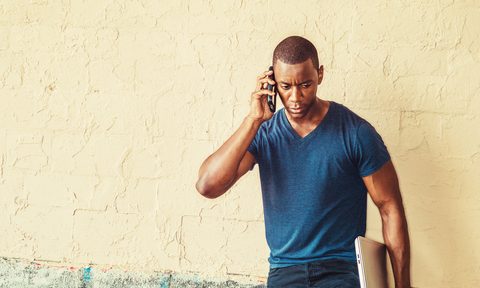 Young handsome man talking on cell phone outdoor, wearing blue V neck T shirt, holding laptop computer, standing against wall on street, looking down, listening.