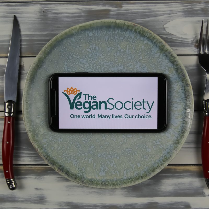 The logo of The Vegan Society displayed on a phone on a plate, with a knife and fork beside it on a table.