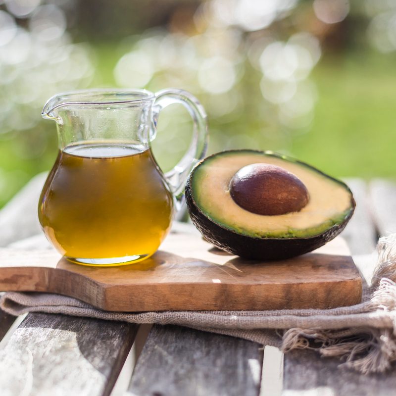 A pitcher of avocado oil beside a sliced avocado on a wooden board.