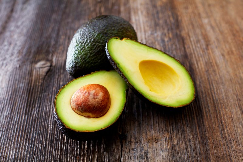 A whole avocado behind a sliced avocado on a wooden surface.