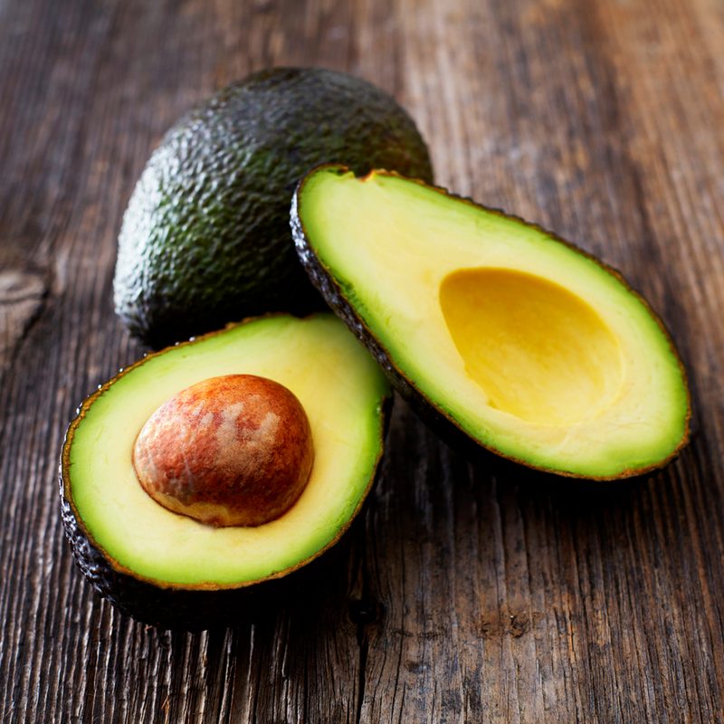 A whole avocado behind a sliced avocado on a wooden surface