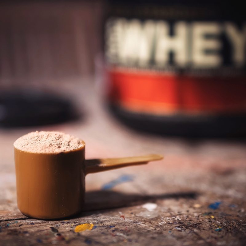 A scoop of protein powder with a jar of protein powder in the background.