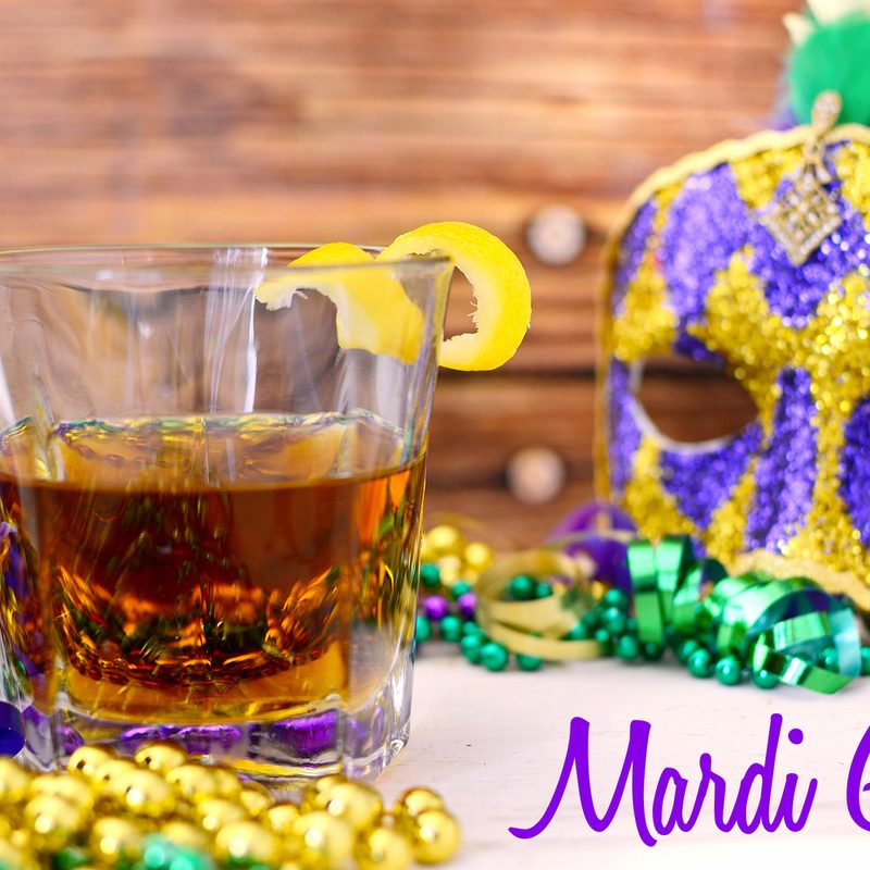 A glass of cocktail beside a mask and gold, purple, and green streamers and beads on wooden background, with “Mardi Gras” at the bottom right corner.