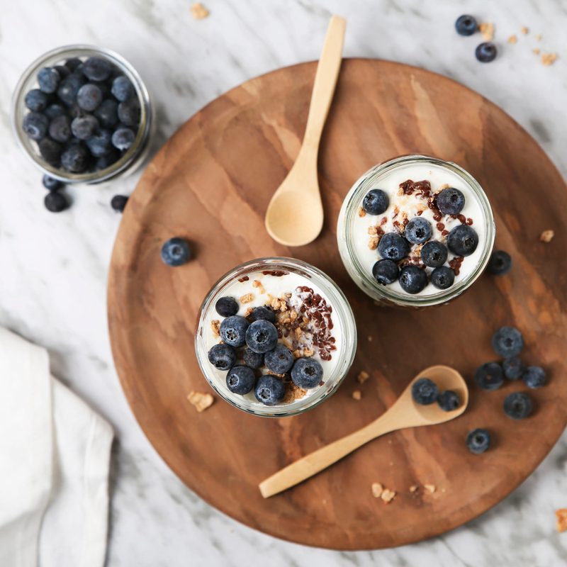 Two cups of yogurt parfait and blueberries on a circular tray beside a bowl of blueberries on a table.