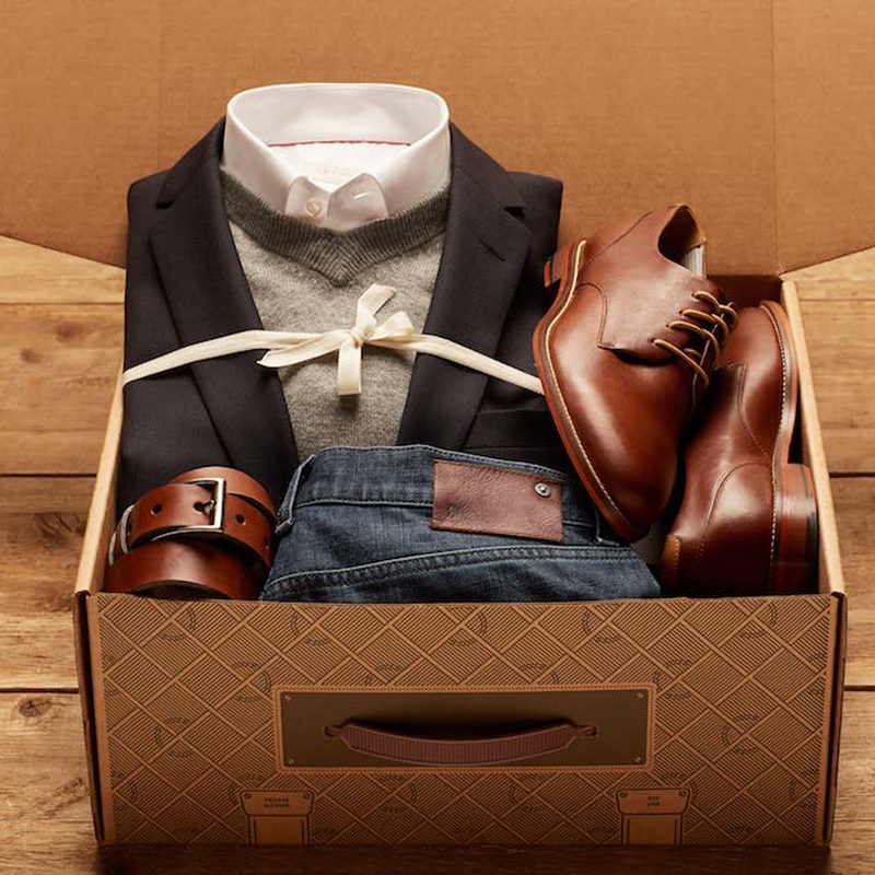 A suit, jeans, belt, and dress shoes in a Trunk Club box on wooden floor.