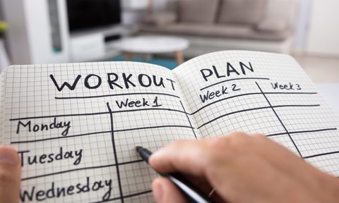 A person's hand writing out their workout plan on a notebook in a room.