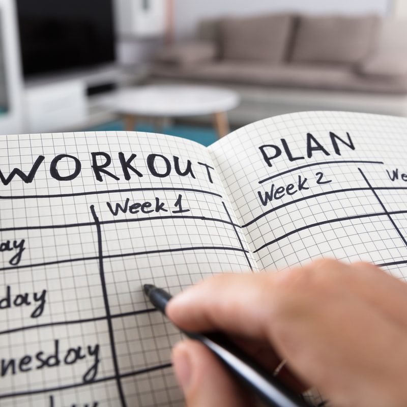 A person's hand writing out their workout plan on a notebook in a room.