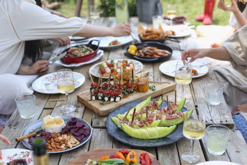 People eating food with wine on a wooden table outside.